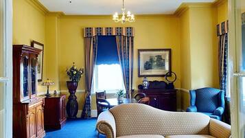 A room with a patterned sofa, a wooden coffee table, a chandelier, a dining table with chairs, and a large window with curtains.