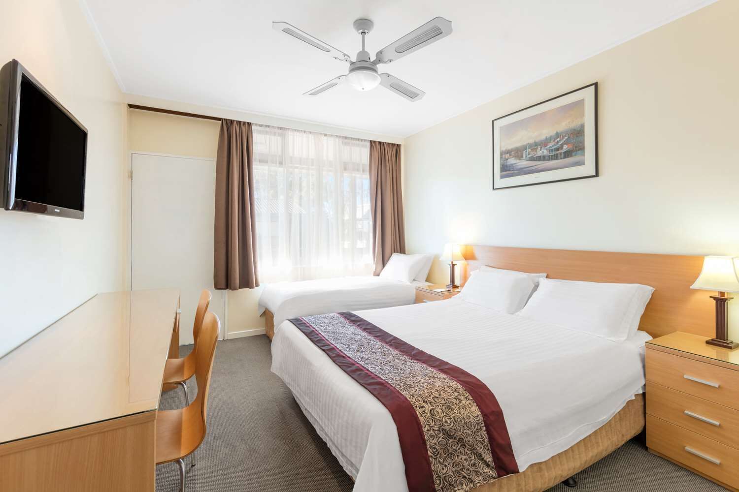 A hotel room with a bed, two bedside tables, a desk, a chair, a ceiling fan, a TV, and a framed picture on the wall.