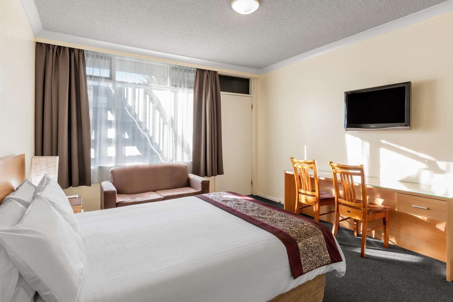 A hotel room with a bed, a desk, two chairs, a television, and a window with curtains.