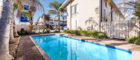 A modern building with a swimming pool, palm trees, and a fenced patio area.