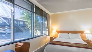 A hotel room with a bed, two bedside lamps, a desk, and a window with a view of palm trees and a building.