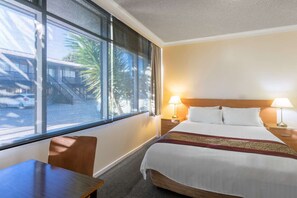 A hotel room with a bed, two bedside lamps, a desk, and a window with a view of palm trees and a building.