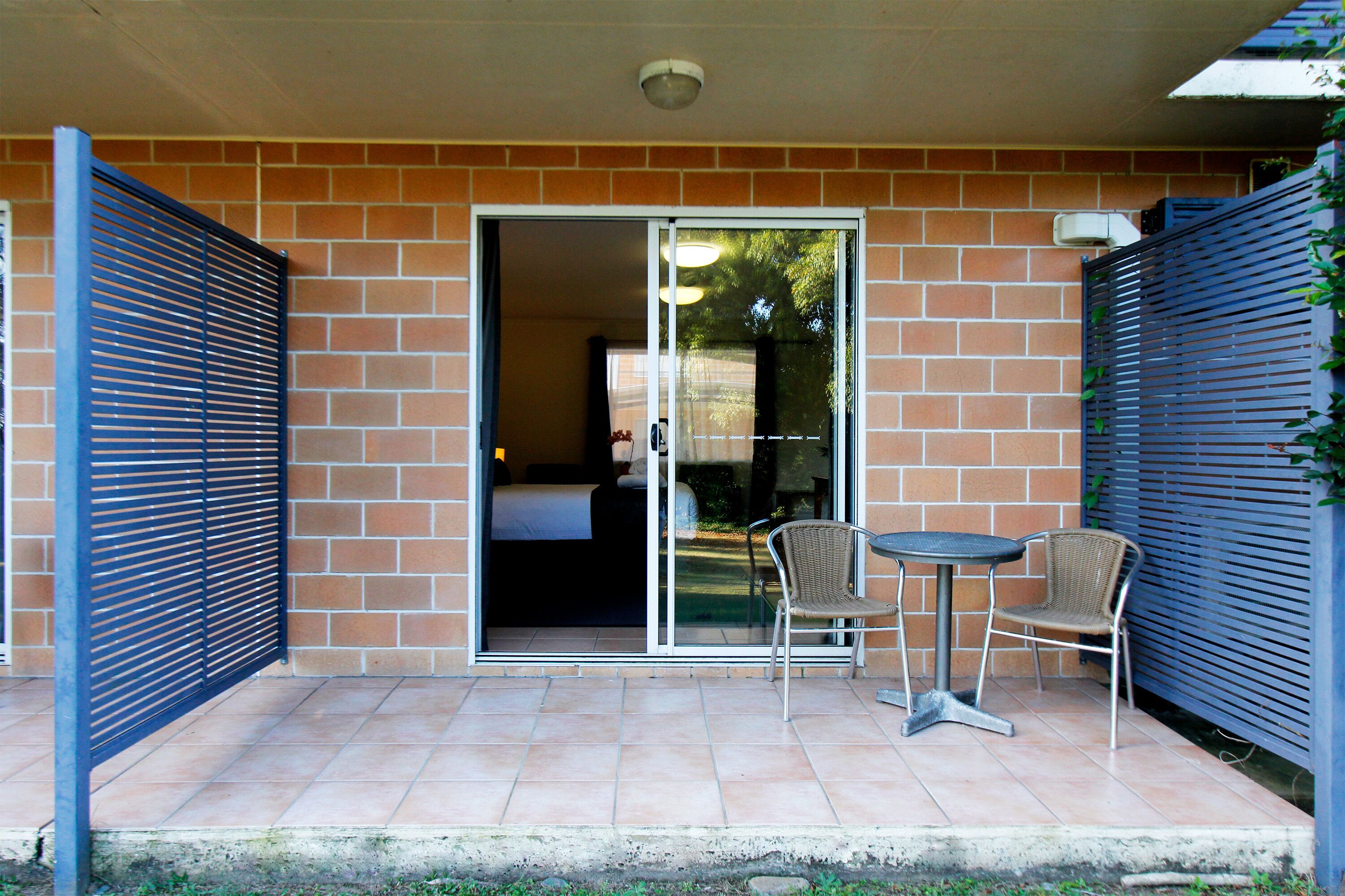 A patio with two chairs and a table, a sliding glass door leading to a room, and a brick wall.