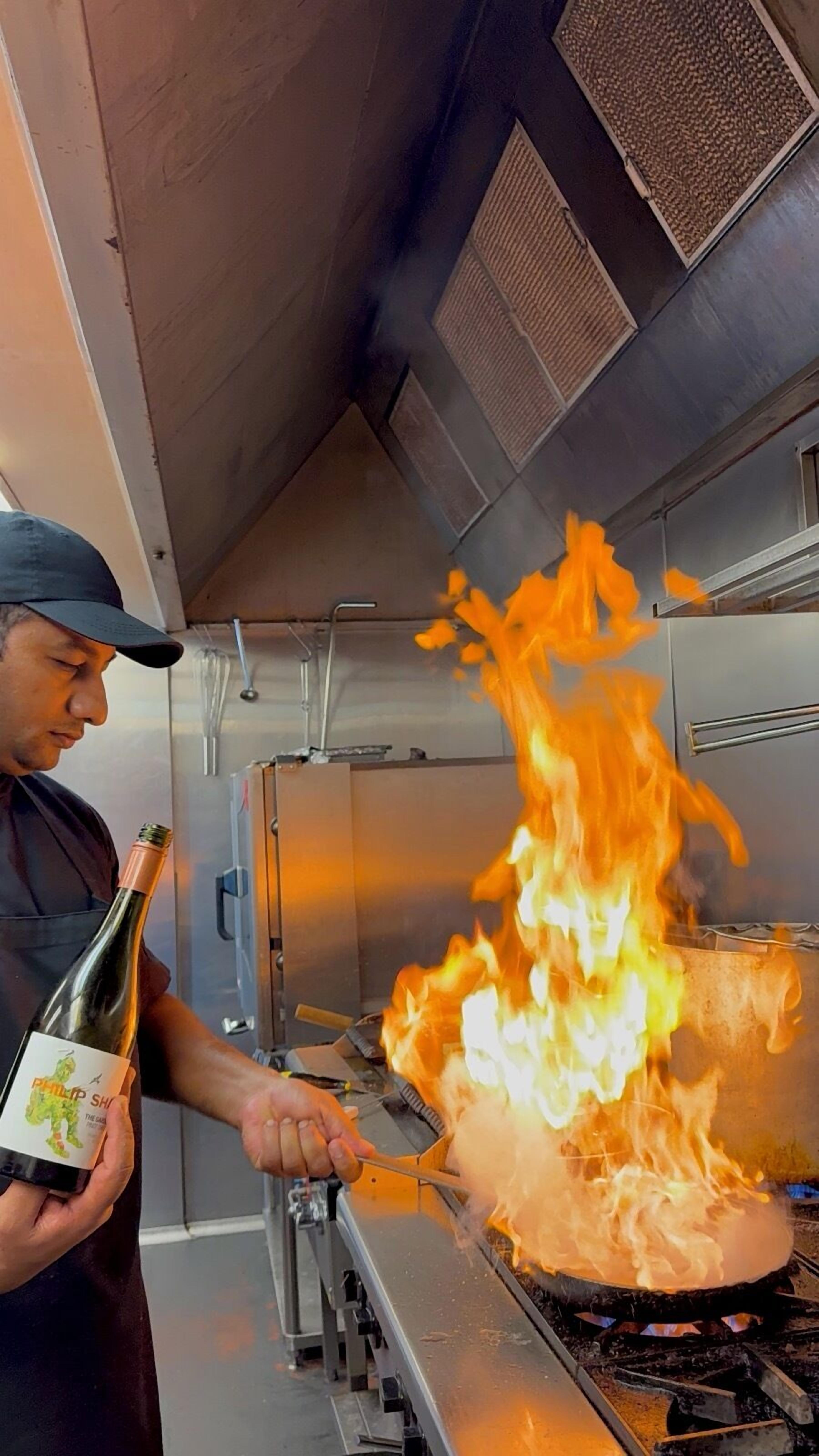 Un chef prépare un plat flambé à l’aide d’une bouteille de vin dans une cuisine professionnelle.