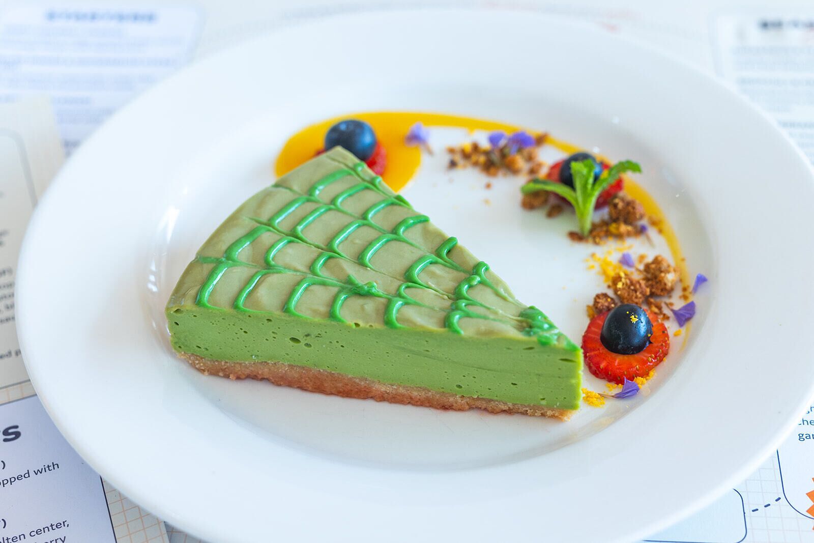 A slice of green dessert on a white plate with a grid pattern on top, accompanied by blueberries and a small dollop of cream.