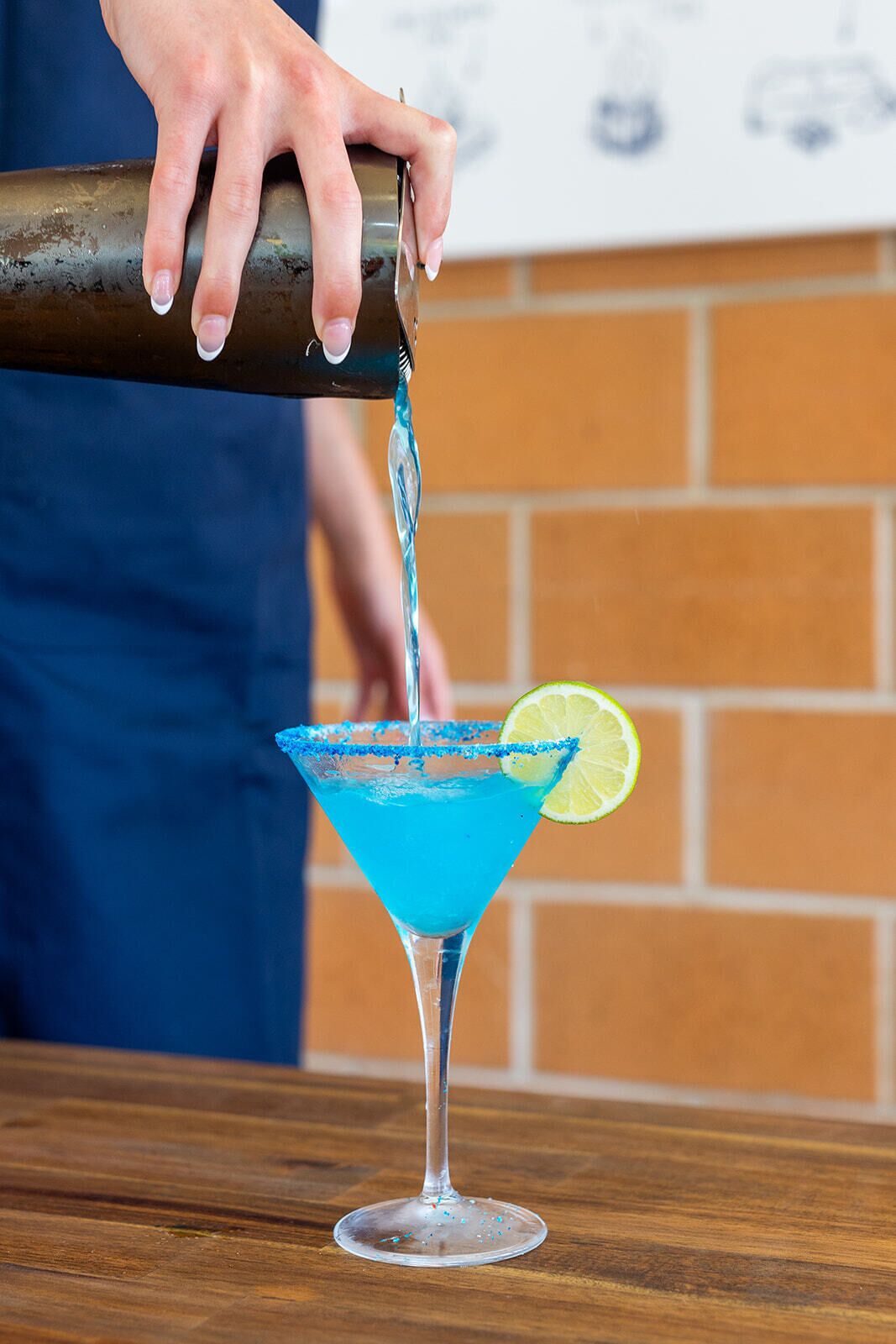 A person pouring a blue liquid into a martini glass with a lime garnish.
