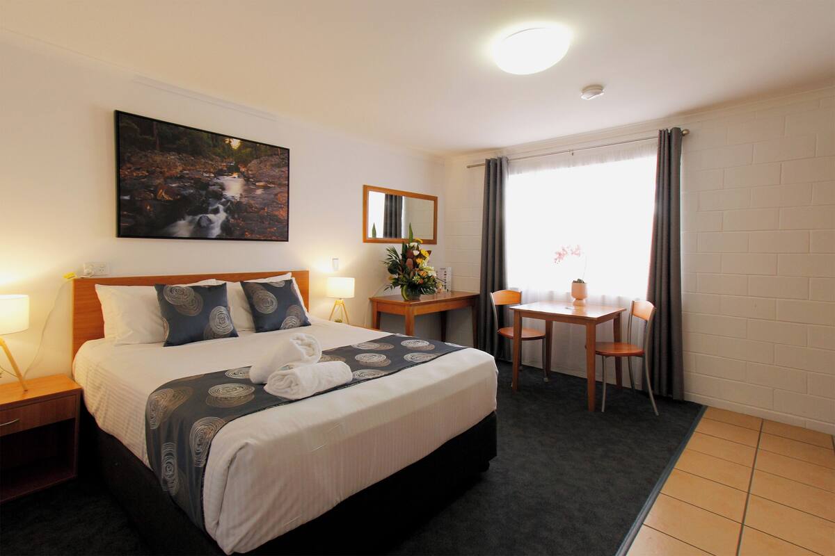 Une chambre d’hôtel avec un grand lit, un bureau, une chaise, une petite table avec un vase, un miroir et un tableau au mur.