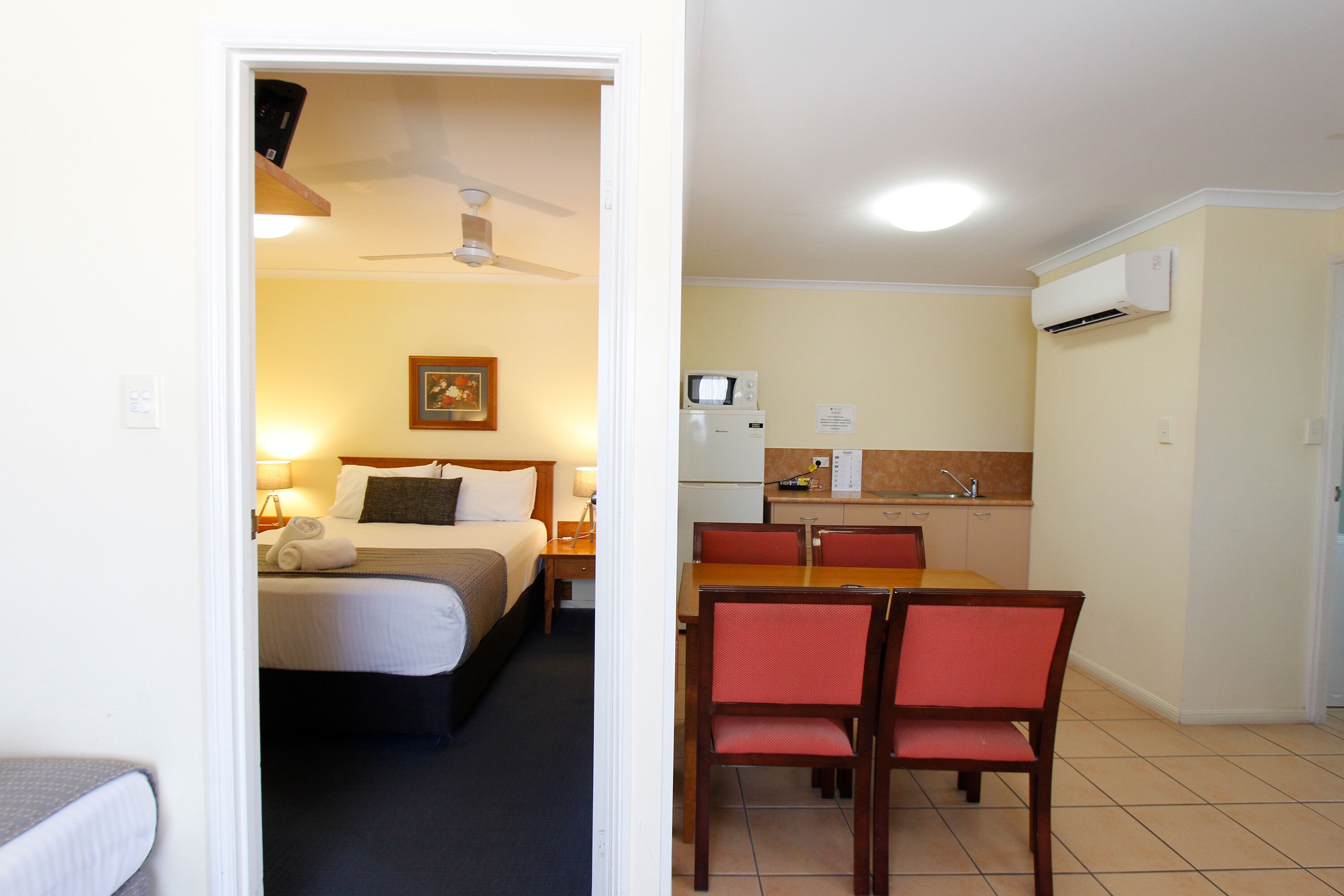 A hotel room with a bed, bedside table, lamp, and a ceiling fan. Adjacent is a kitchenette with a microwave, sink, and a small dining table with chairs.
