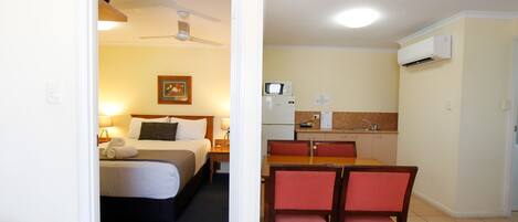 Habitación de hotel con cama, mesita de noche, lámpara y ventilador de techo. Junto a ella, una kitchenette con microondas, fregadero y una pequeña mesa de comedor con sillas.