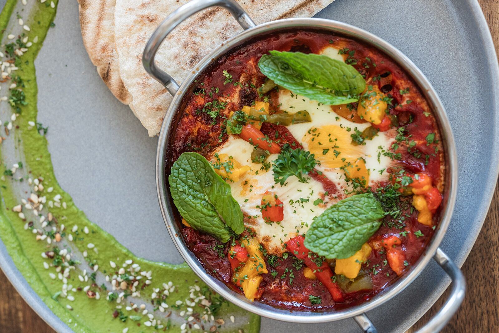 A bowl of stew with vegetables and cheese, garnished with herbs, served with flatbread.