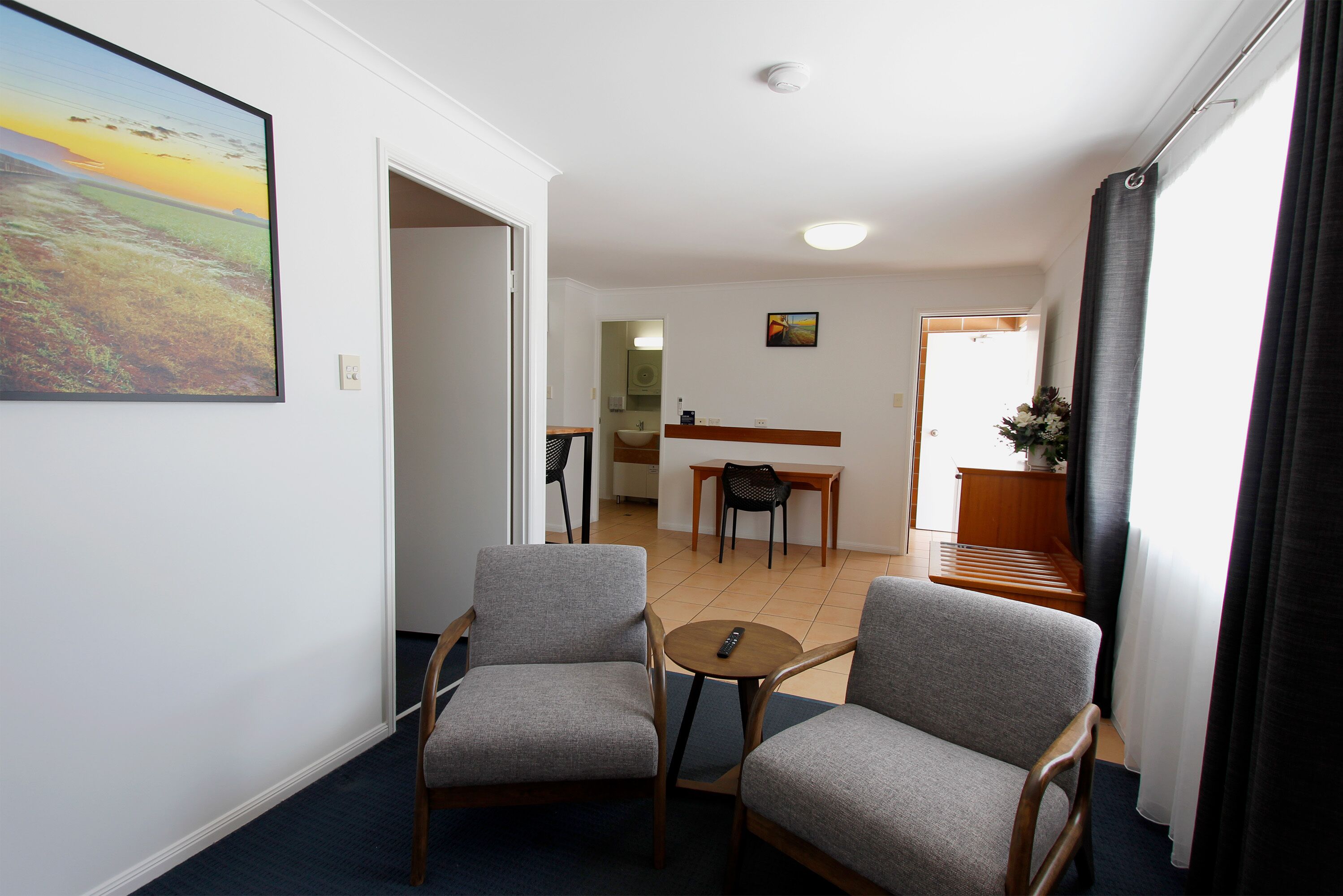 A room with two grey armchairs, a small round table, a painting on the wall, a kitchen area in the background, and a staircase leading to another level.