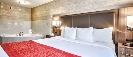 Habitación de hotel con una cama grande, una manta roja, un cabecero de madera, una mesita de noche con lámpara y un baño con lavamanos y espejo.
