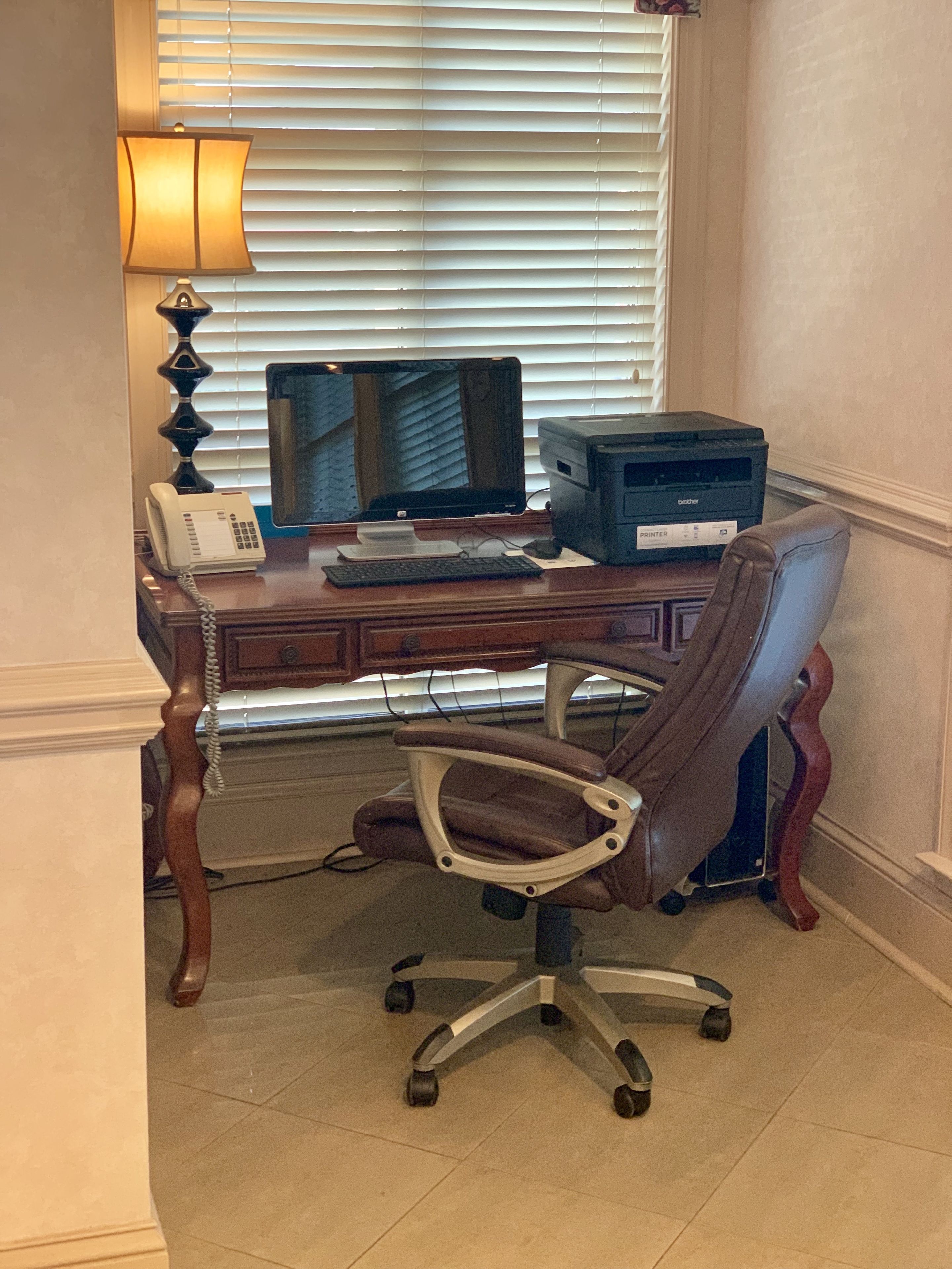 A wooden desk with a computer monitor, printer, and telephone. A brown office chair is in front of the desk.