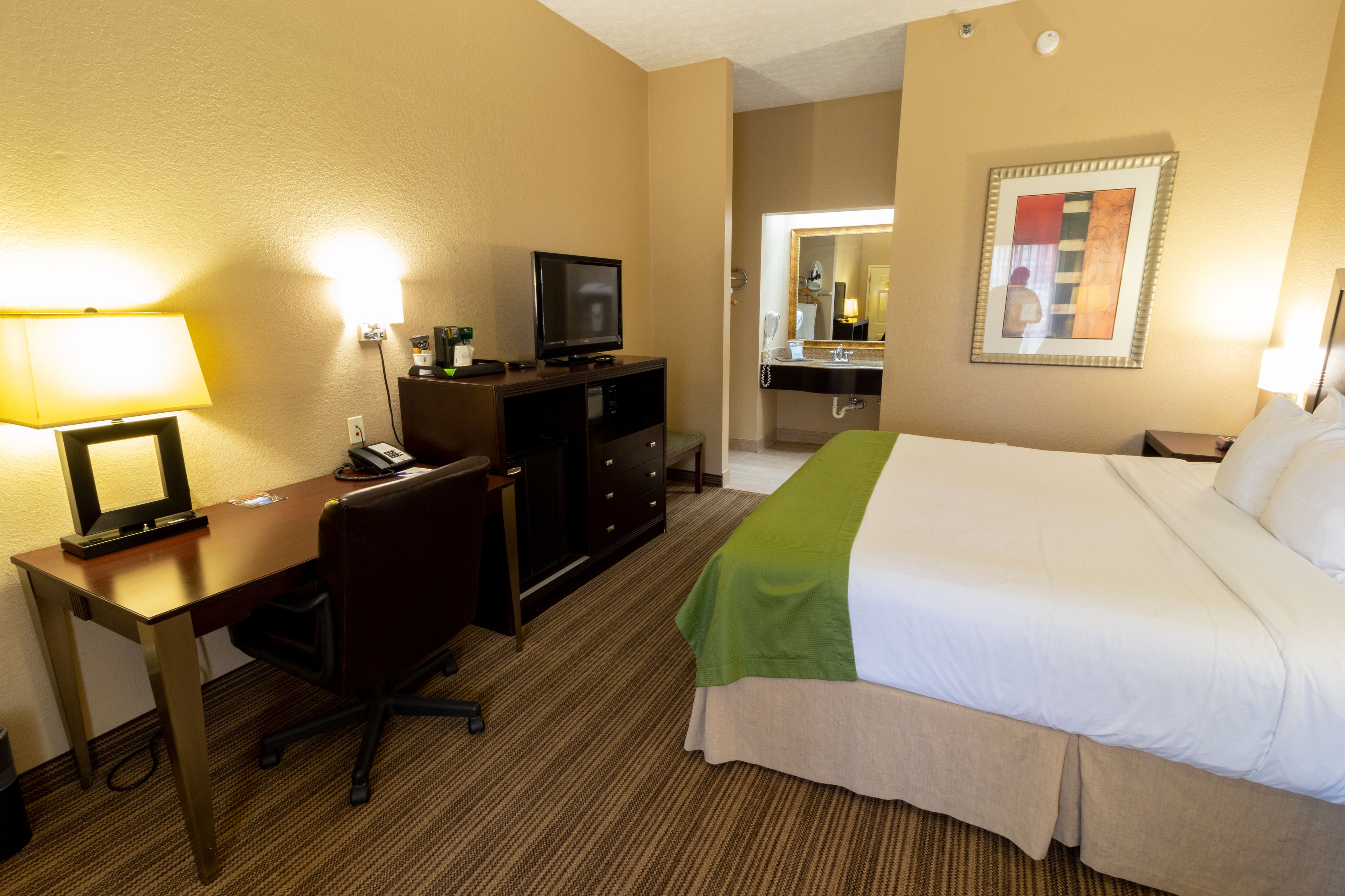 Habitación de hotel con cama, escritorio, silla, televisión y baño con espejo y lavamanos.