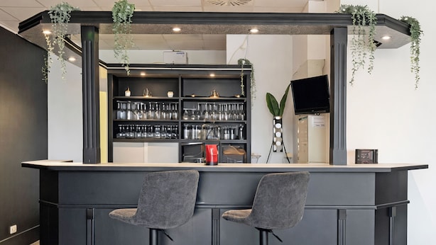 A modern bar area with a dark counter, two grey bar stools, and shelves stocked with glasses and bottles. - DORMERO Hotel Bretten (Bretten)