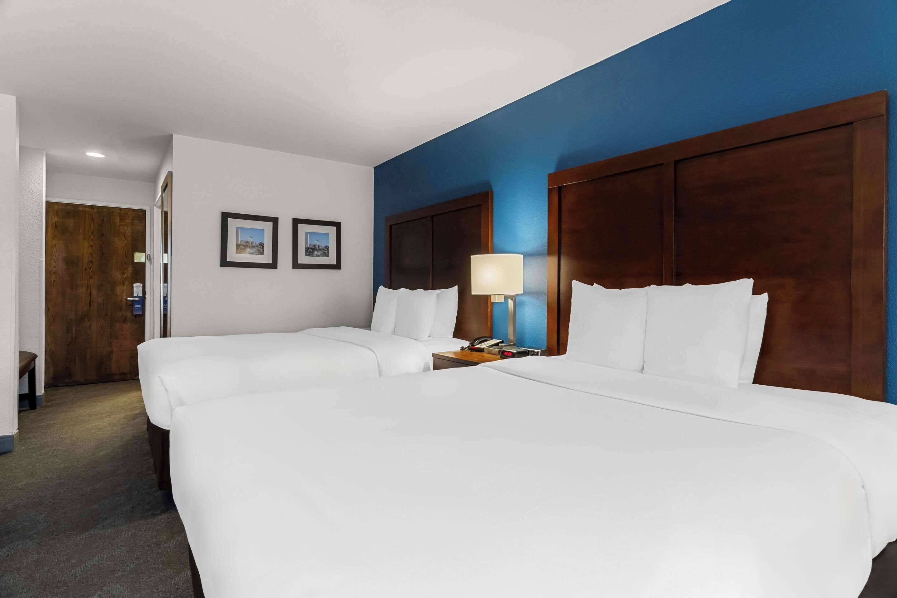 A hotel room with two beds, a wooden headboard, a nightstand with a lamp, and two framed pictures on the wall.