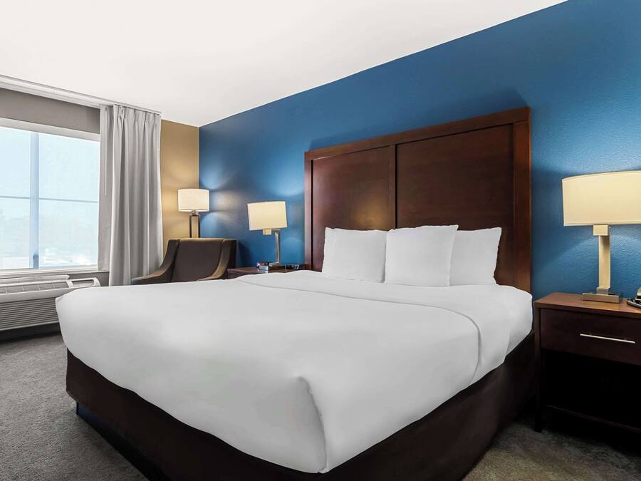 Una habitación de hotel con una cama grande, dos mesitas de noche con lámparas, una silla y un ventanal con cortinas.