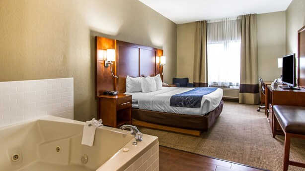 Ett hotellrum med en stor säng, ett separat skrivbord med en stol, en TV, ett badrum med badkar och ett fönster med gardiner.