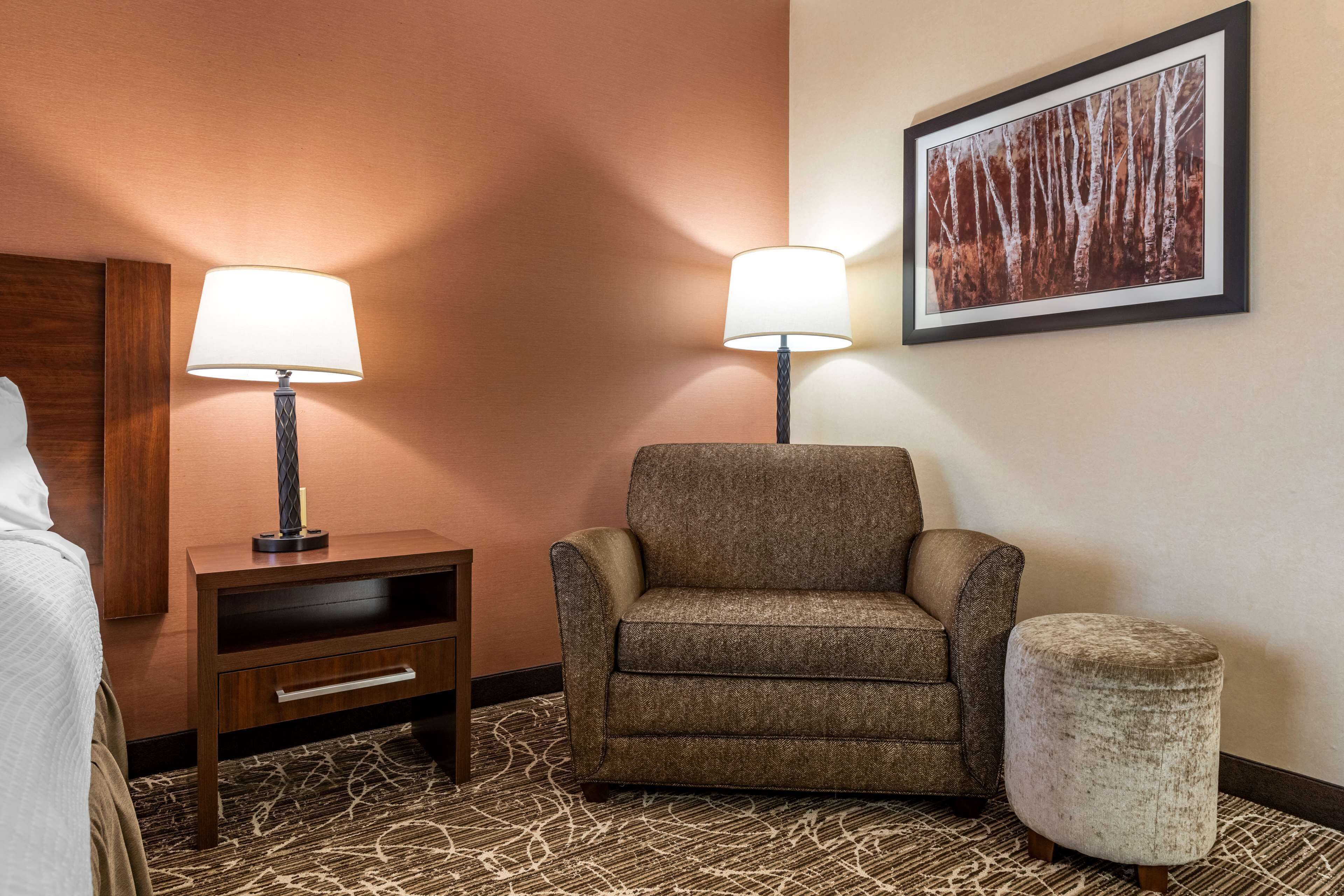 Habitación de hotel con un sillón marrón, un pequeño reposapiés, una mesita de noche con una lámpara y un cuadro enmarcado en la pared.