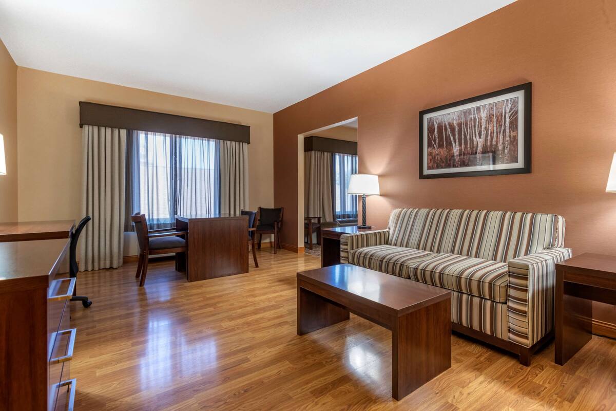 A hotel room with a wooden floor, a striped sofa, a desk with a chair, a lamp, and a framed picture on the wall.