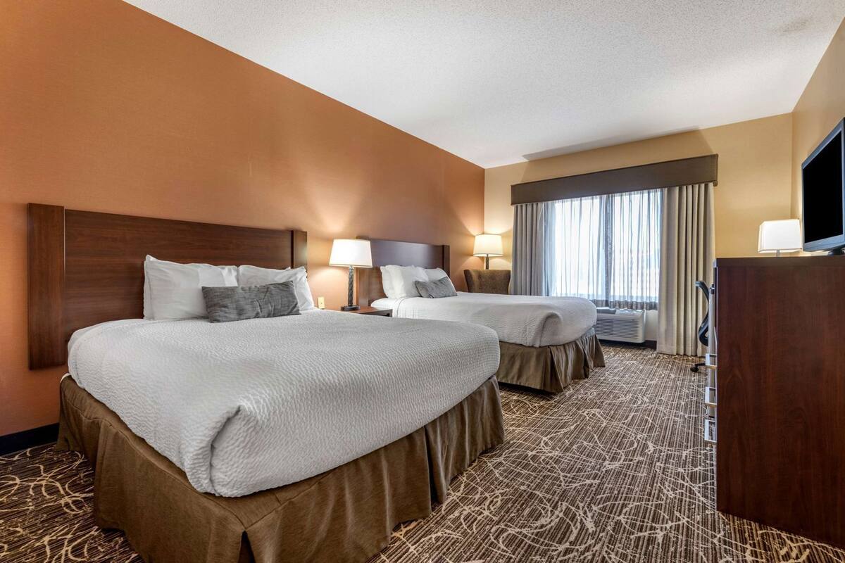 A hotel room with two beds, a TV, a desk, and a large window with curtains.