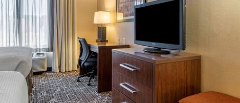 Una habitación de hotel con cama, escritorio con silla, televisión, lámpara y un cuadro en la pared.
