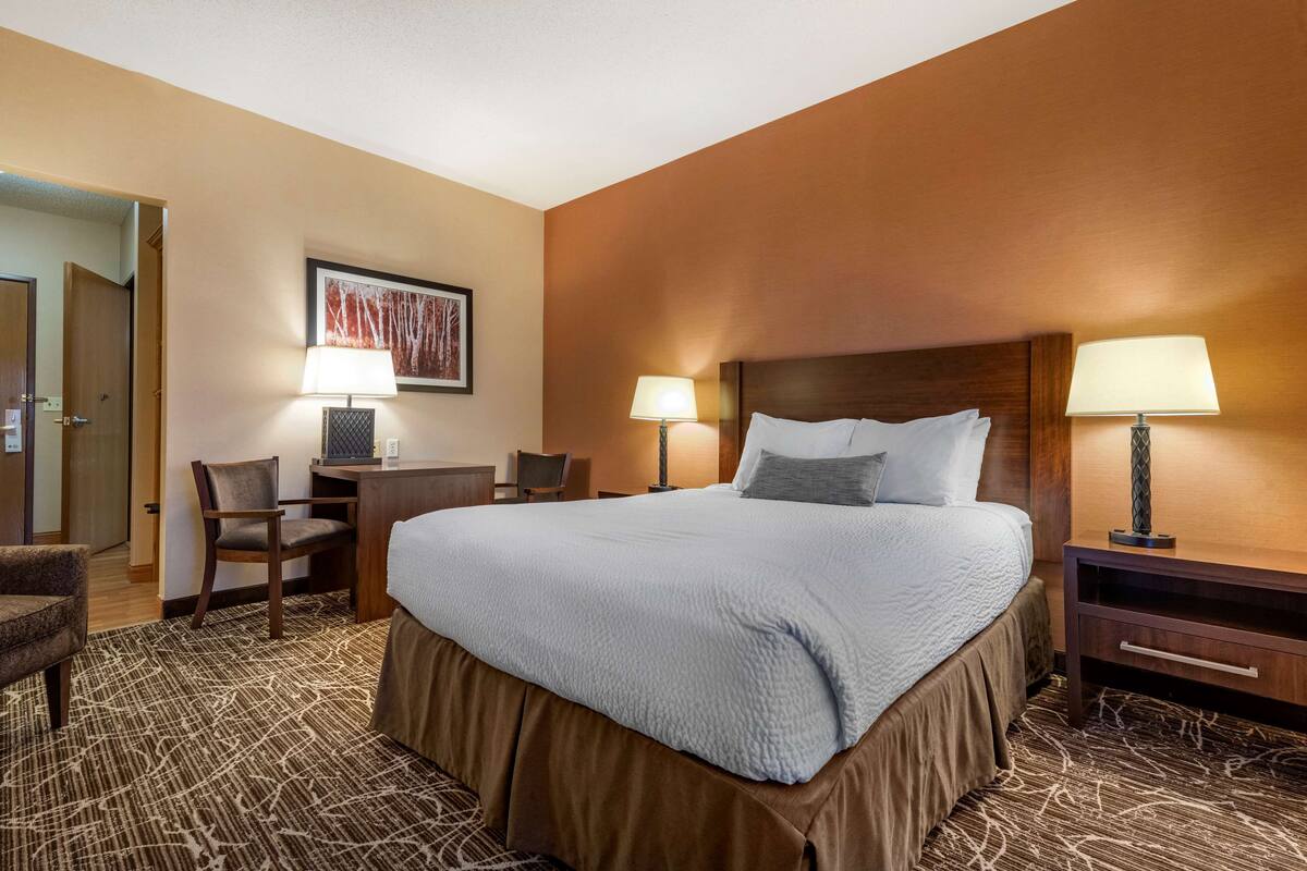 A hotel room with a large bed, two bedside tables with lamps, a chair, a desk, and a framed picture on the wall.