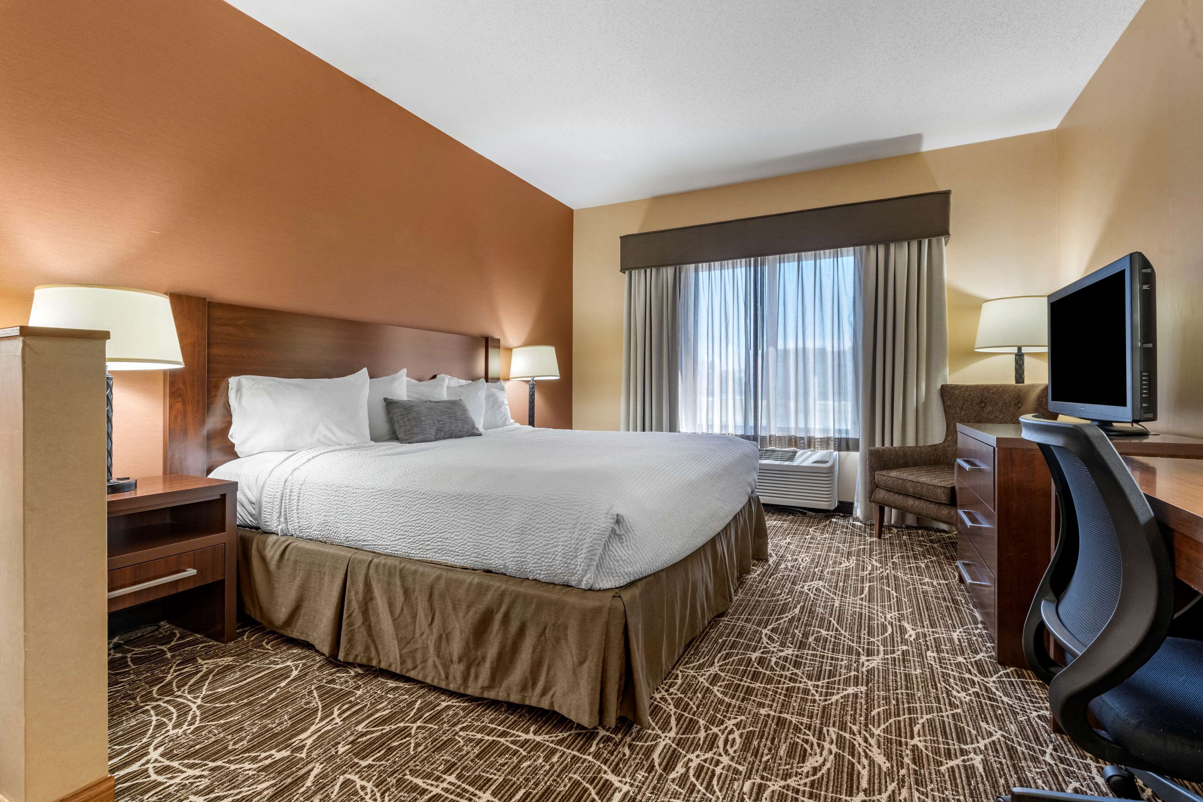 A hotel room with a large bed, a desk with a computer, a TV, and a window with curtains.