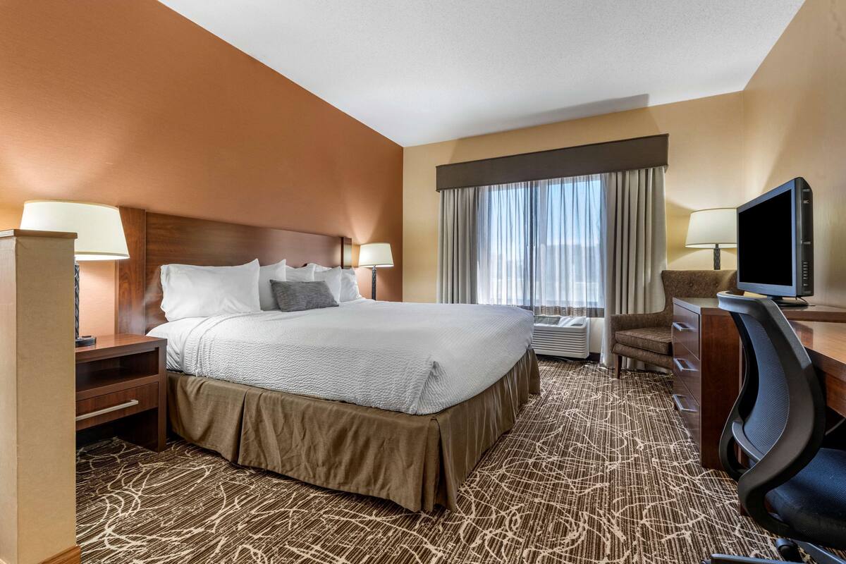 A hotel room with a large bed, a desk with a computer, a TV, and a window with curtains.