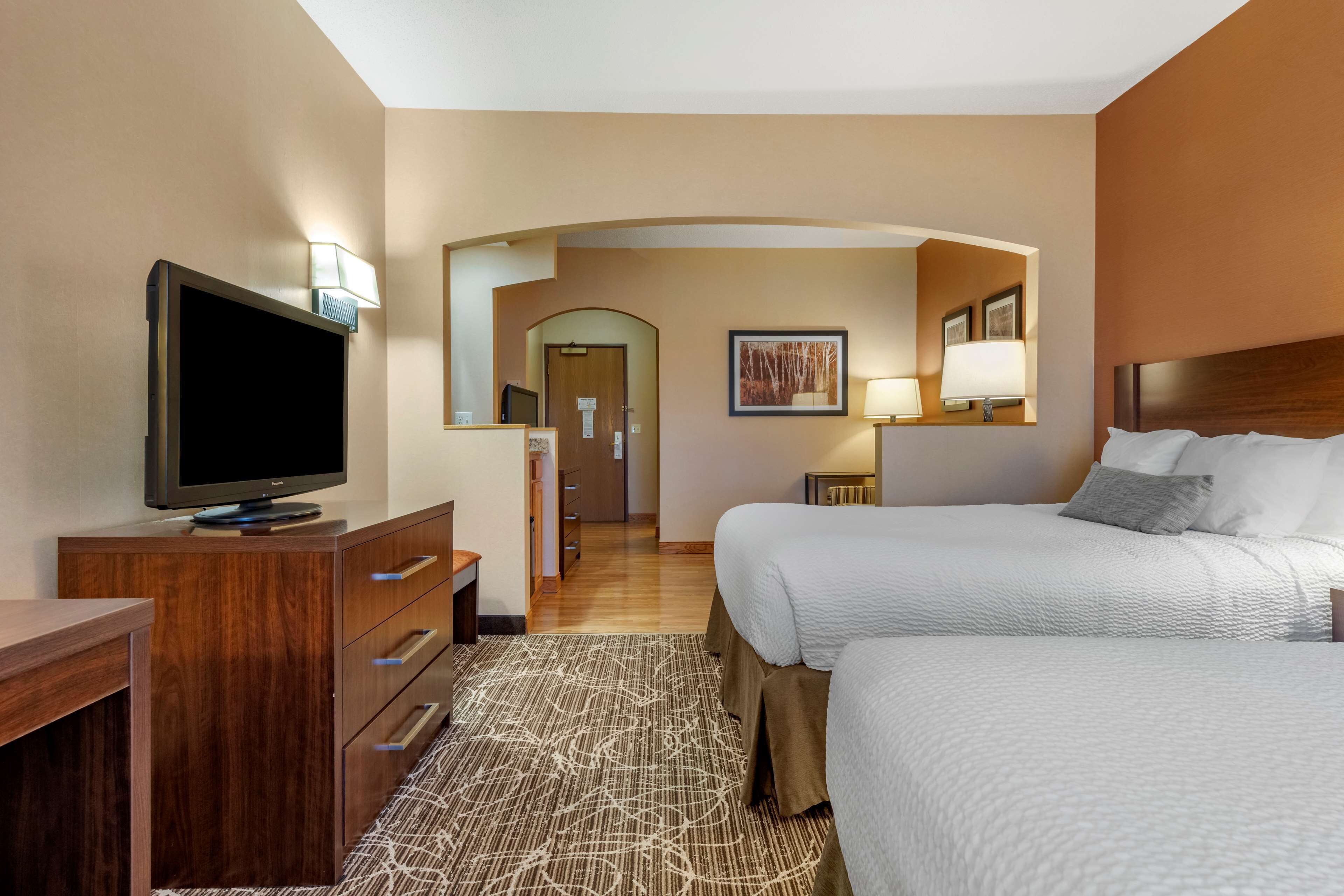 A hotel room with a TV on a wooden dresser, two beds with white bedding, a patterned rug, and a hallway with a mirror and framed picture.