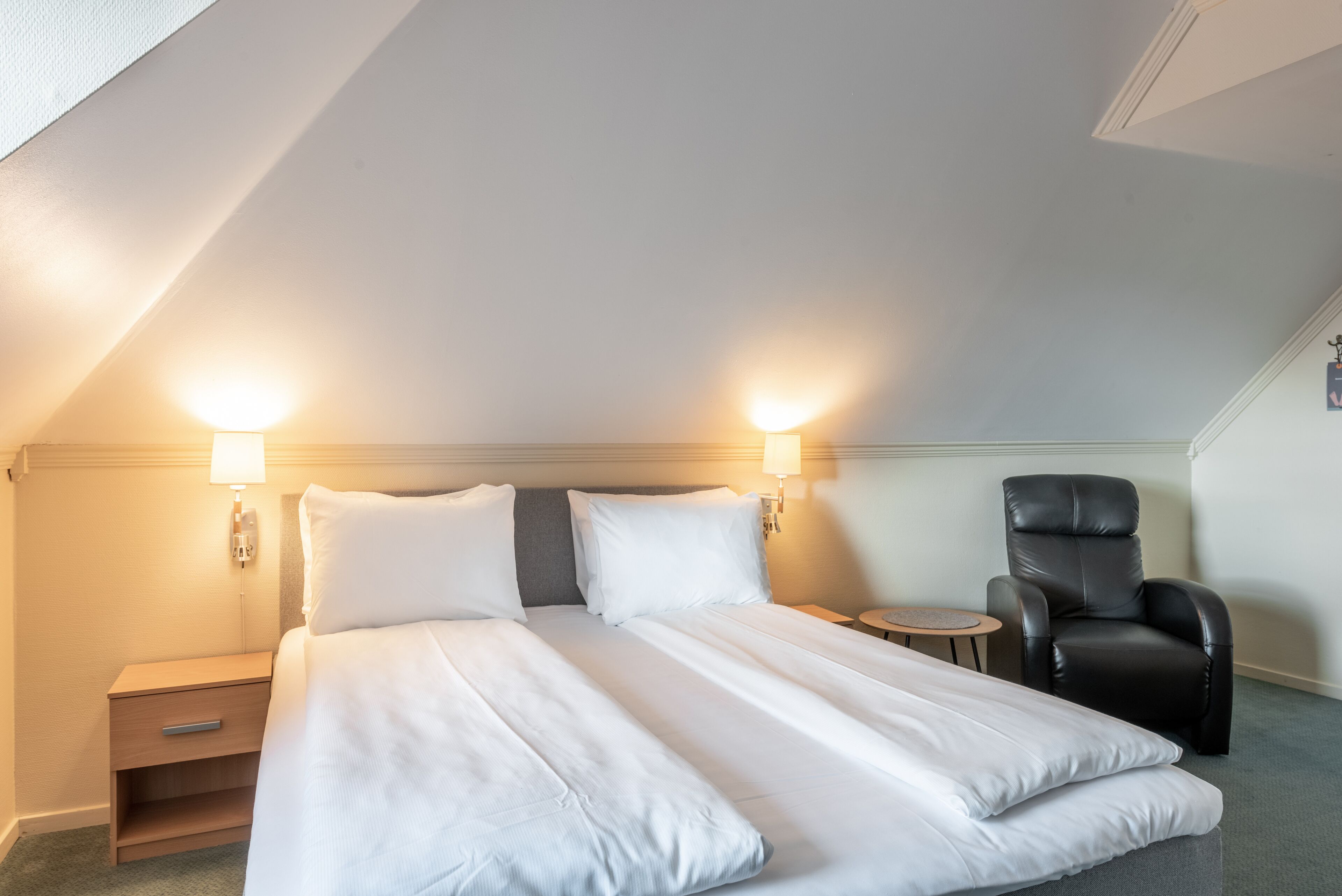 A hotel room with a large bed, two bedside lamps, a small round table, and a leather chair.