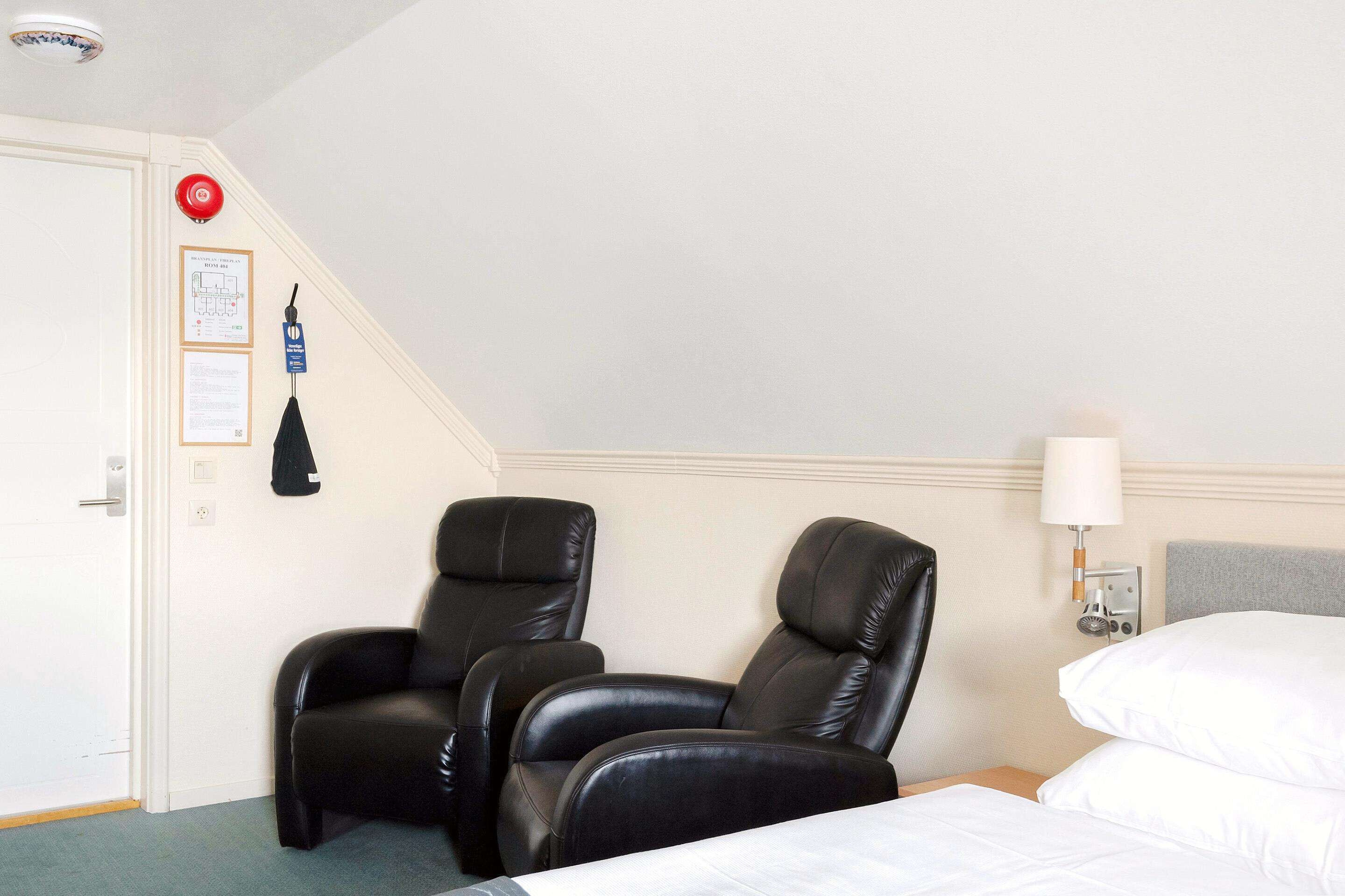 Habitación de hotel con una cama, dos sillones de masaje negros, una lámpara de pared y una puerta con alarma de emergencia roja.