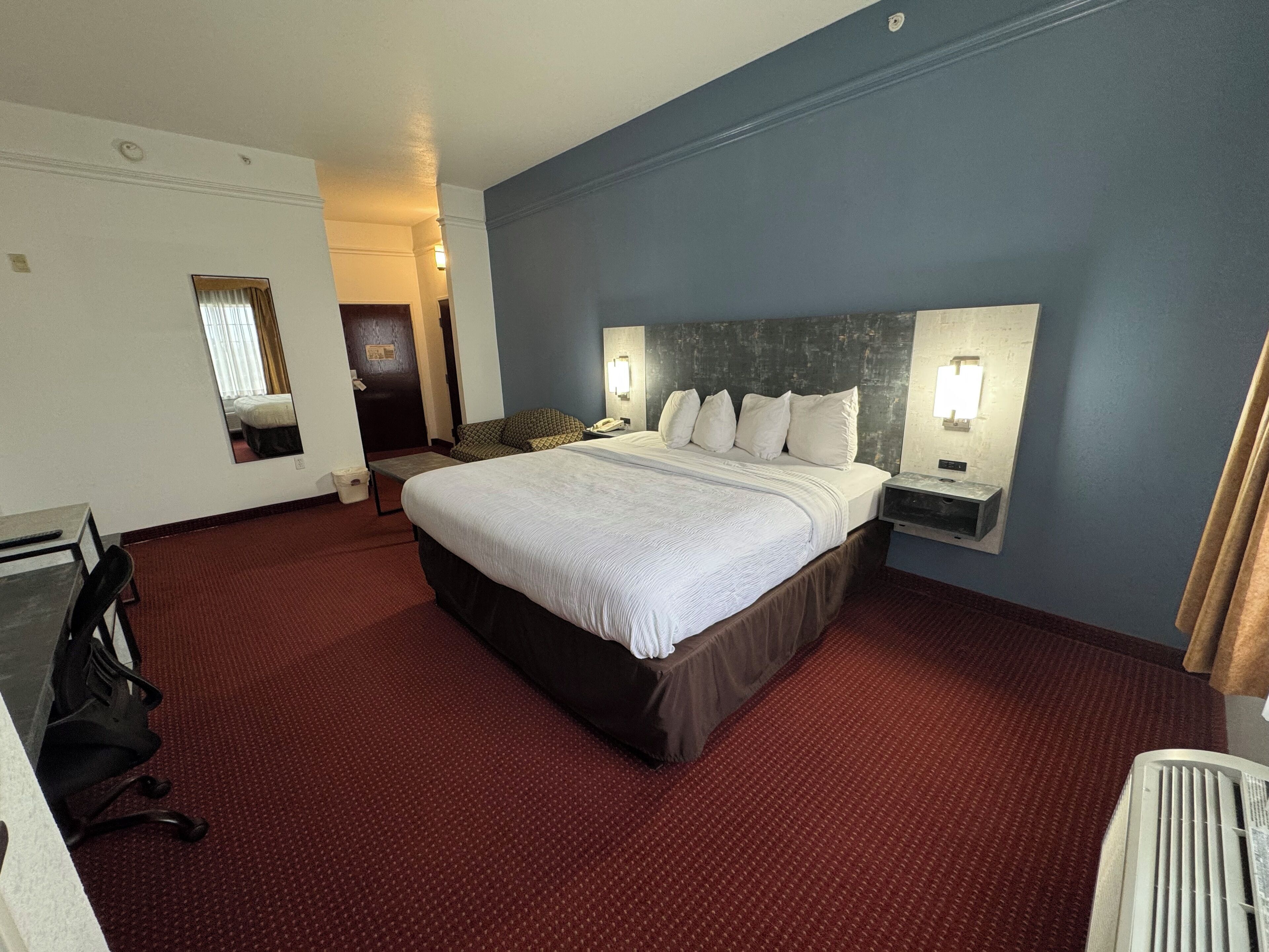 Habitación de hotel con una cama grande, dos mesitas de noche con lámparas, un escritorio con silla y un espejo en la pared.