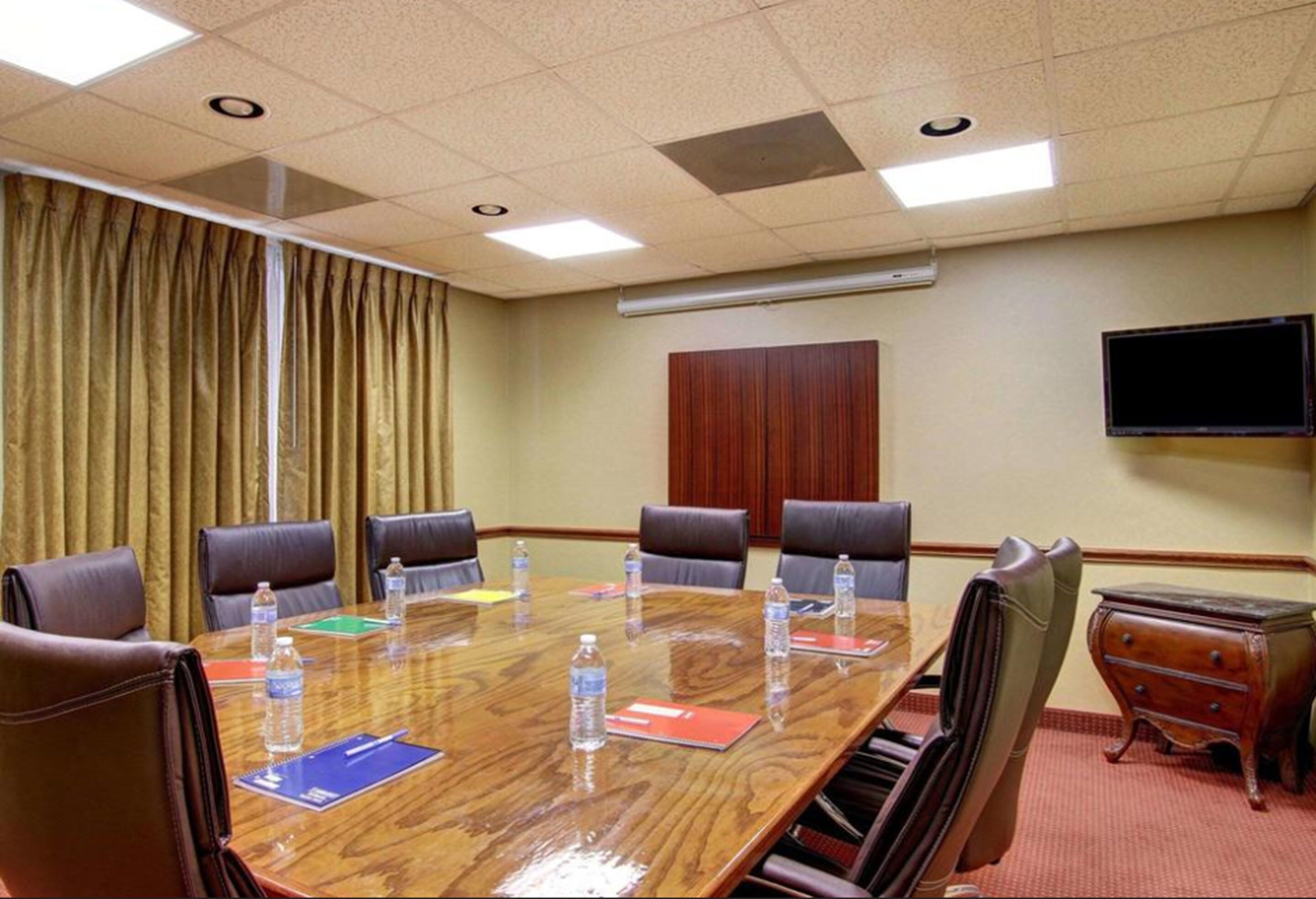 Un salón de conferencias con una mesa de madera, sillas de cuero, botellas de agua, tarjetas de identificación y una televisión en la pared.