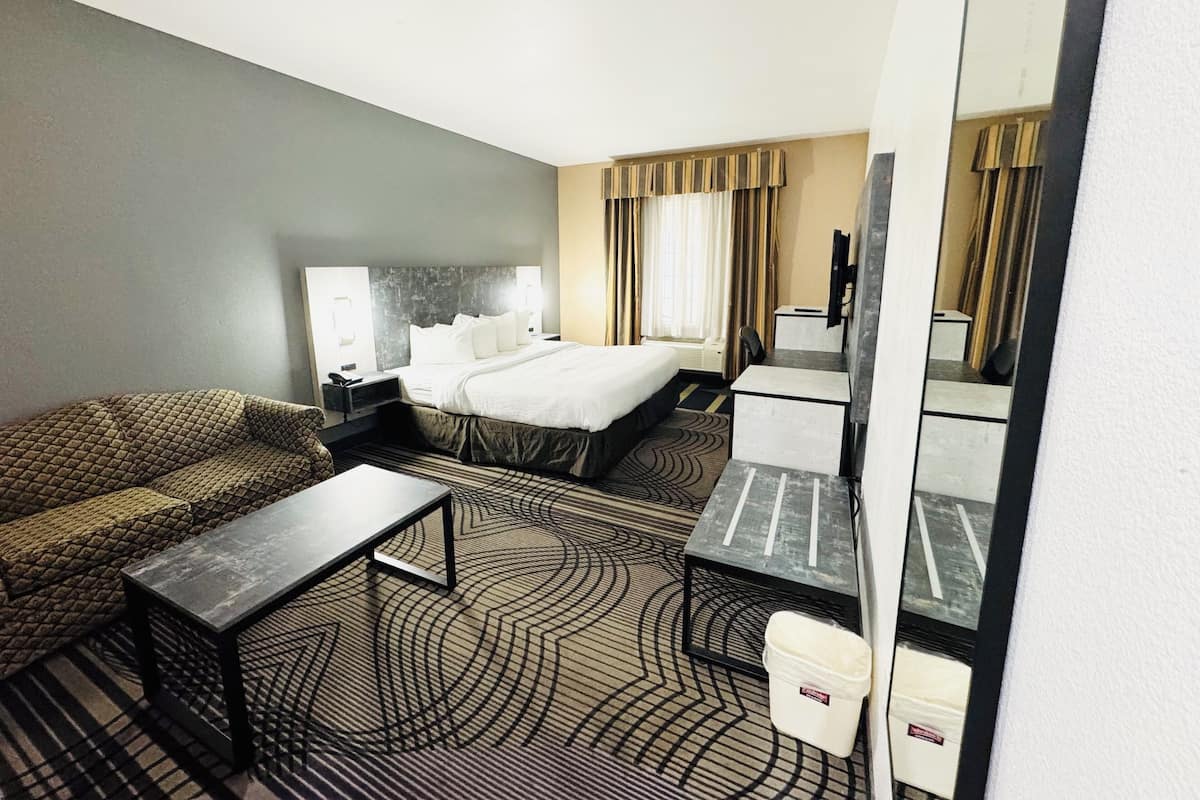 Quarto de hotel com cama, sofá, escrivaninha, cadeira, mesa de cabeceira, abajur, espelho e lixeira.