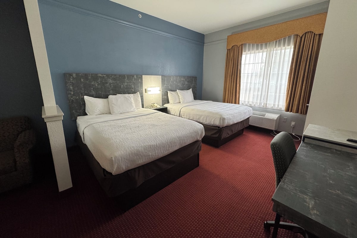Quarto de hotel com duas camas, uma escrivaninha, uma cadeira, uma janela com cortinas e um radiador.