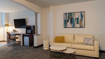 A modern hotel room with a sofa, a small coffee table, a desk with a computer and a flat-screen TV, and a unique abstract painting on the wall.