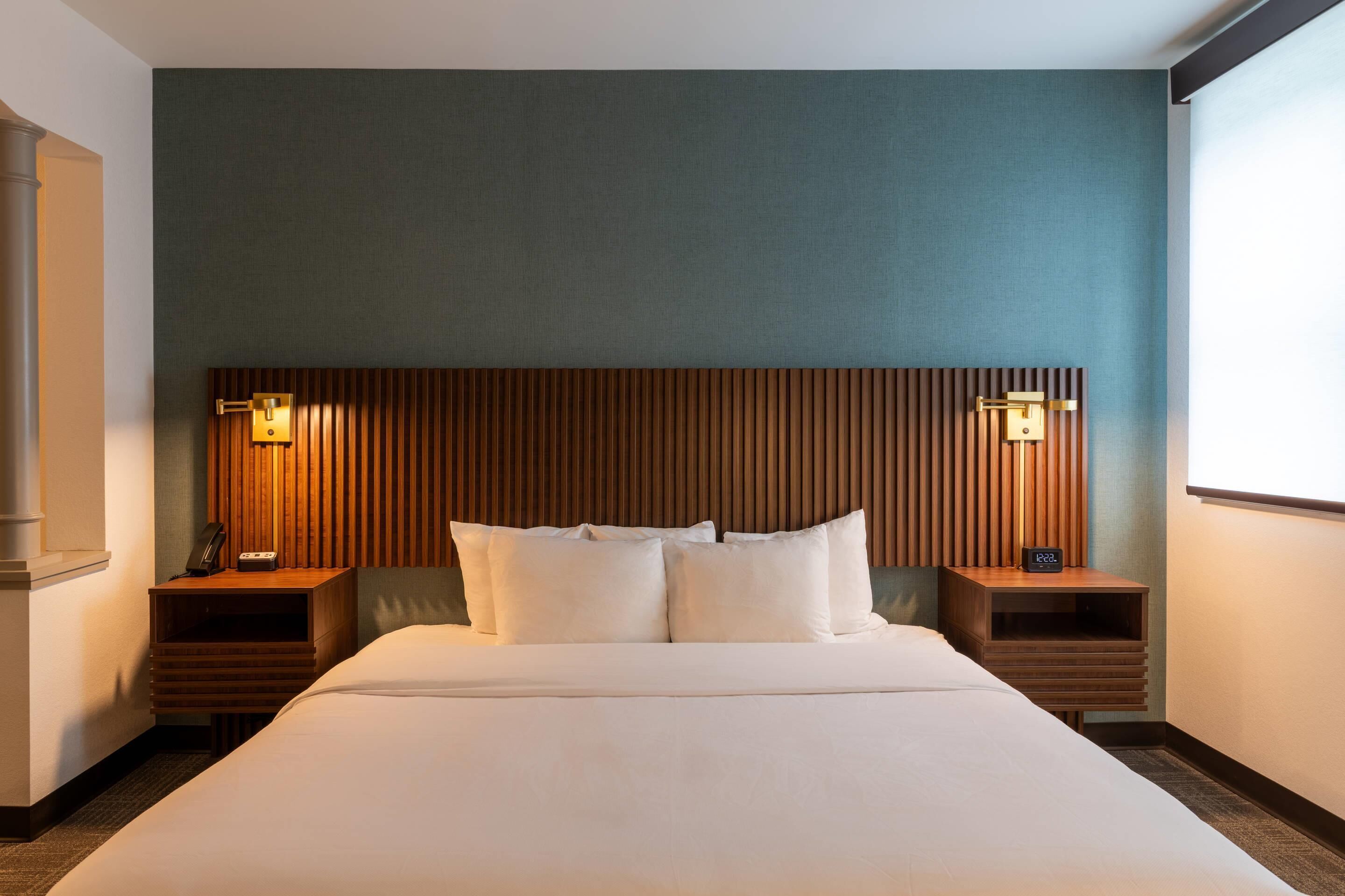A hotel room with a large bed, two bedside tables with lamps, a wooden headboard, and a view of the outdoors.