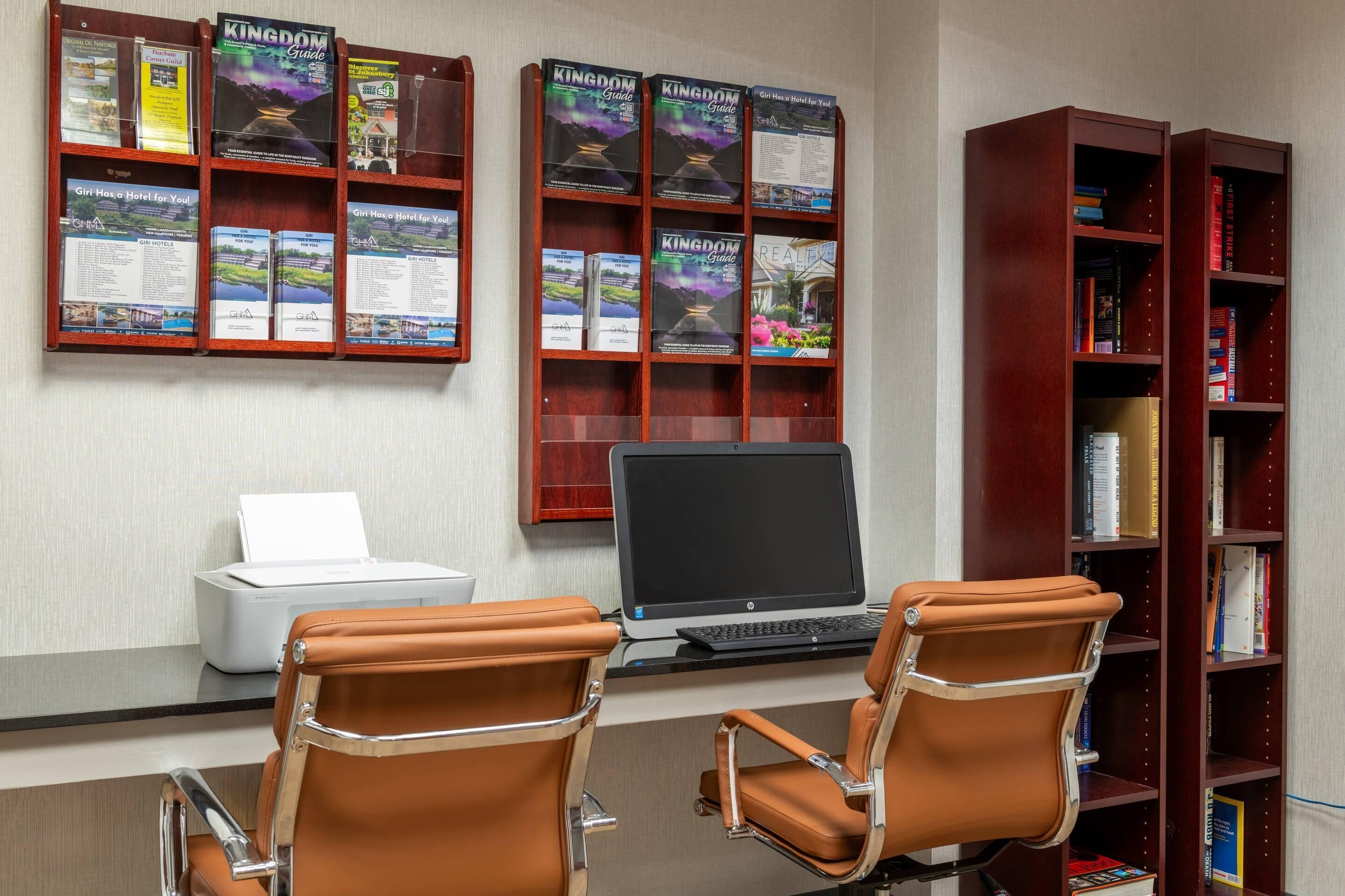 Un mostrador de recepción de hotel con una computadora, dos sillones de cuero, una impresora y una estantería llena de guías de viaje.