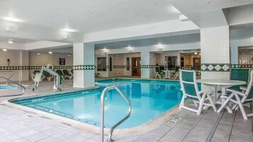Una piscina interior con suelo de baldosas, sillas junto a la piscina y una mesa con sillas.