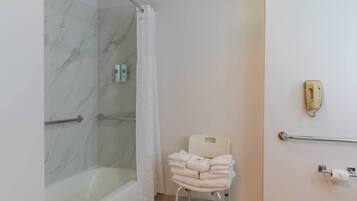 Baño con pared de ducha de mármol, bañera blanca, silla blanca con toallas, barandal y dispensador de papel higiénico.