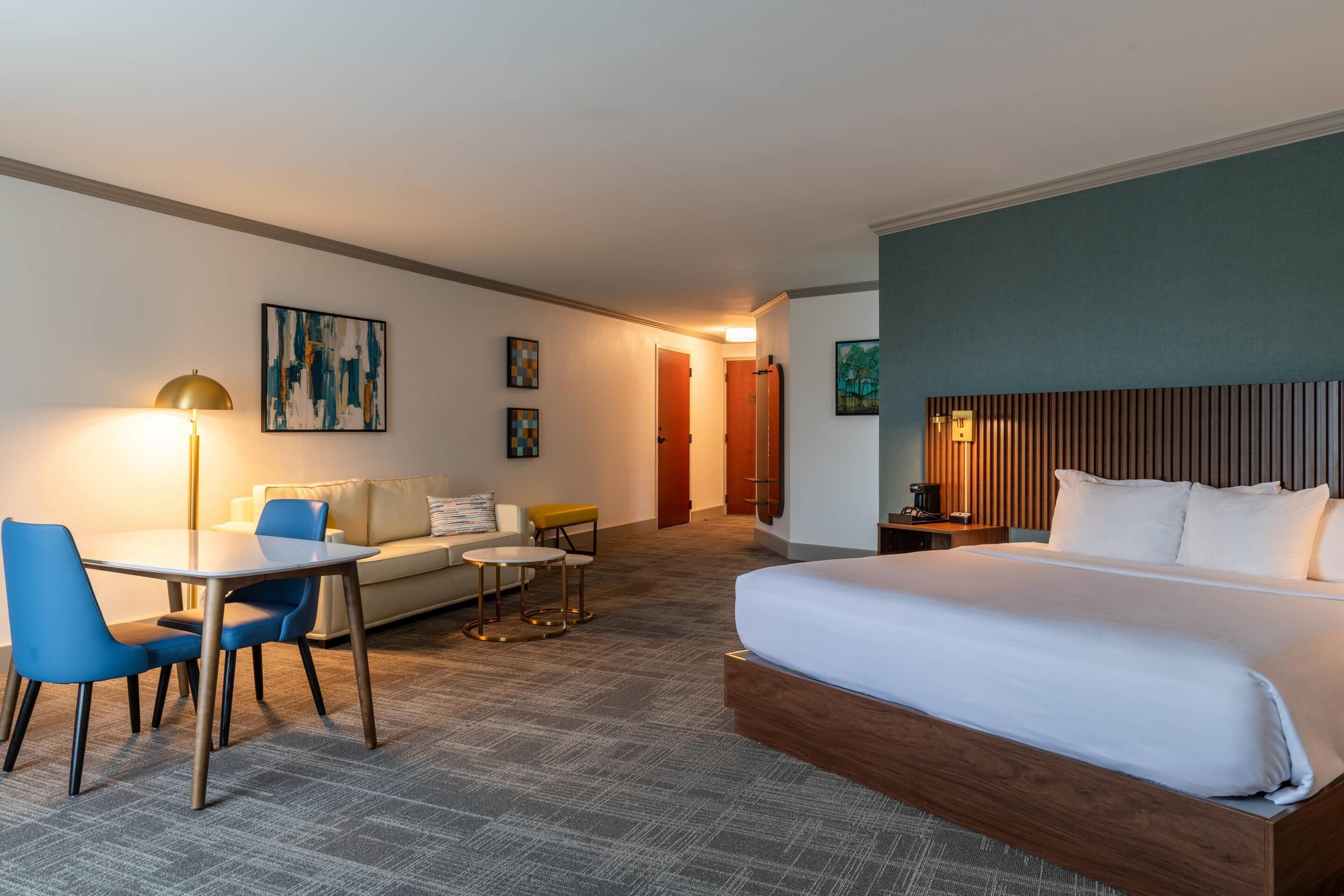 A hotel room with a large bed, a desk with chairs, a sofa, and a small table. There are framed pictures on the walls and a lamp on the desk.