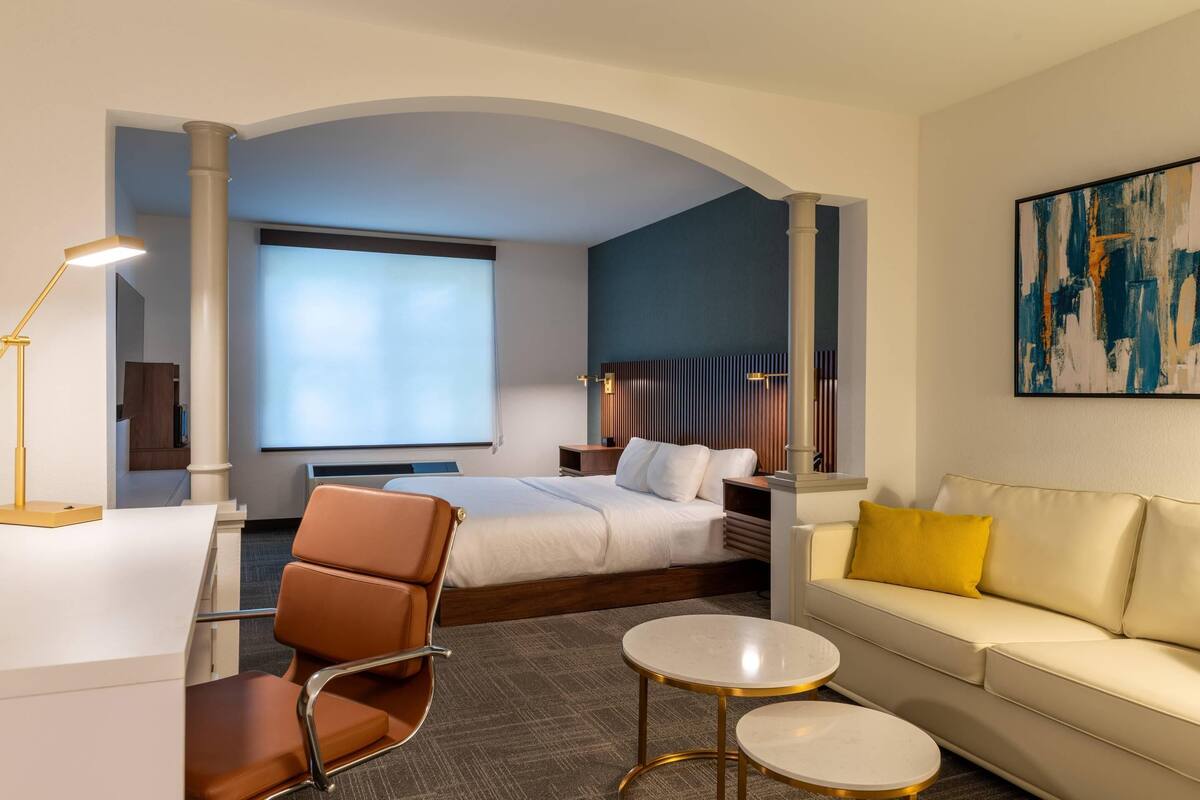 Une chambre d’hôtel avec un lit, un canapé, un bureau et une chaise. Il y a une grande fenêtre et un tableau au mur.
