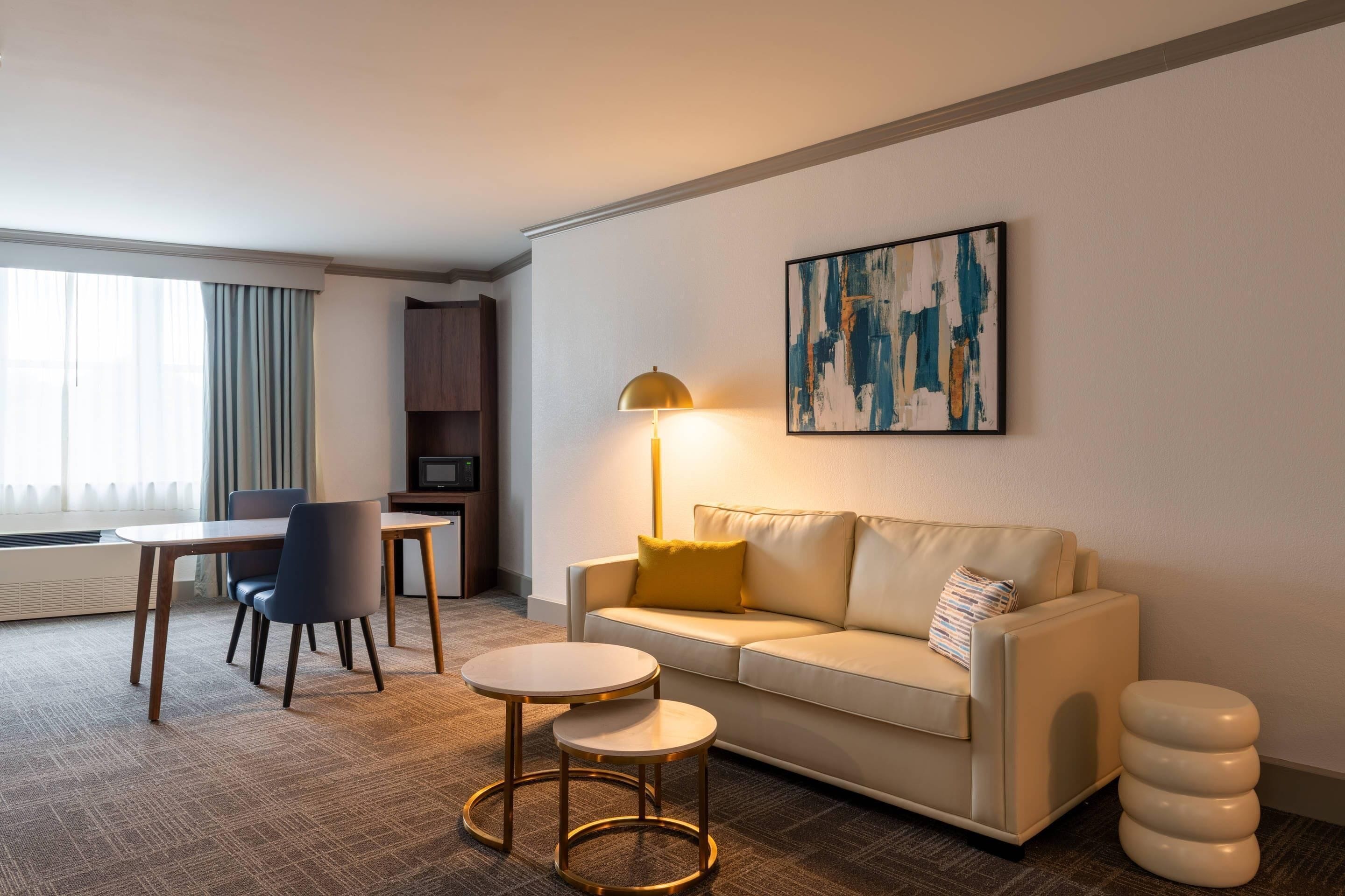 A modern hotel room with a sofa, two round tables, a lamp, and a painting on the wall.