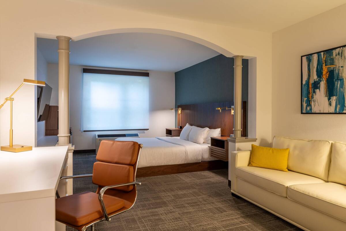 Une chambre d’hôtel avec un lit, un bureau et une chaise, un canapé, une grande fenêtre et un tableau au mur.