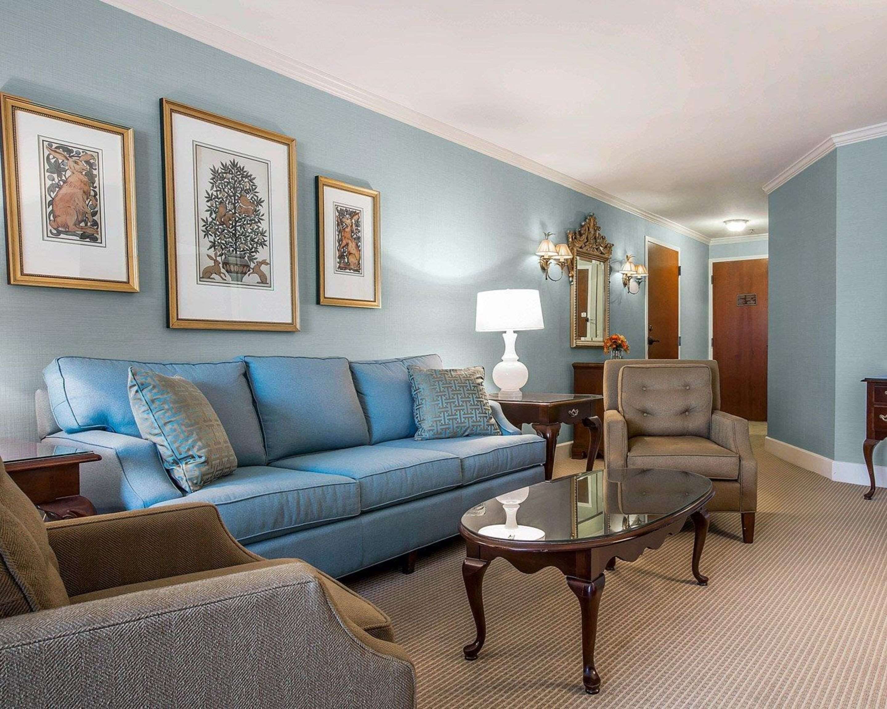 Una sala de estar con un sofá azul, un sillón marrón, una mesa de centro de vidrio y cuadros en la pared.