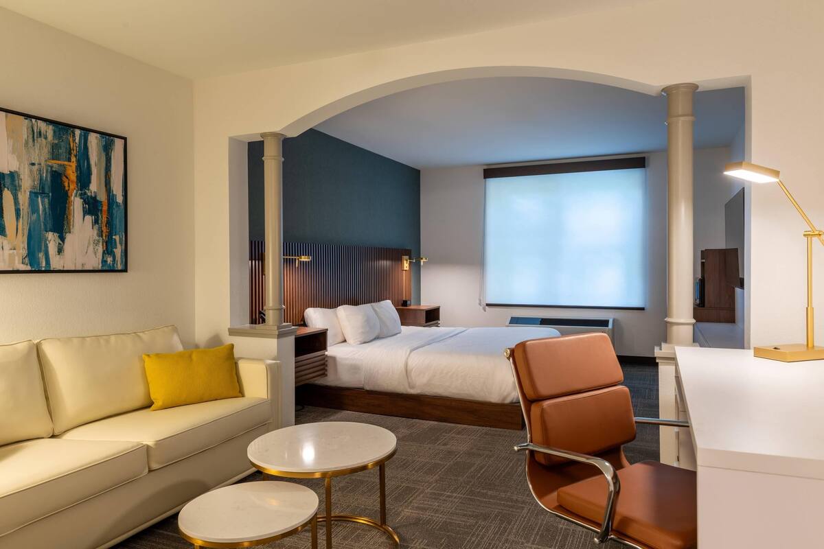 Une chambre d’hôtel avec un lit, un canapé, un bureau et une chaise. Il y a un tableau au mur et une fenêtre avec vue.