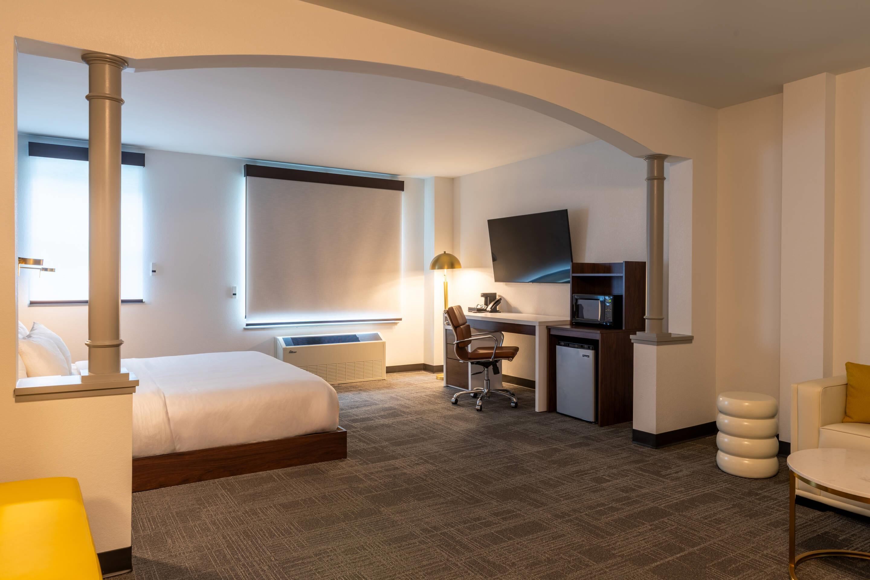 A hotel room with a bed, desk, chair, TV, and a small kitchenette.