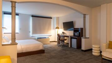 A hotel room with a bed, desk, chair, TV, and a small kitchenette.
