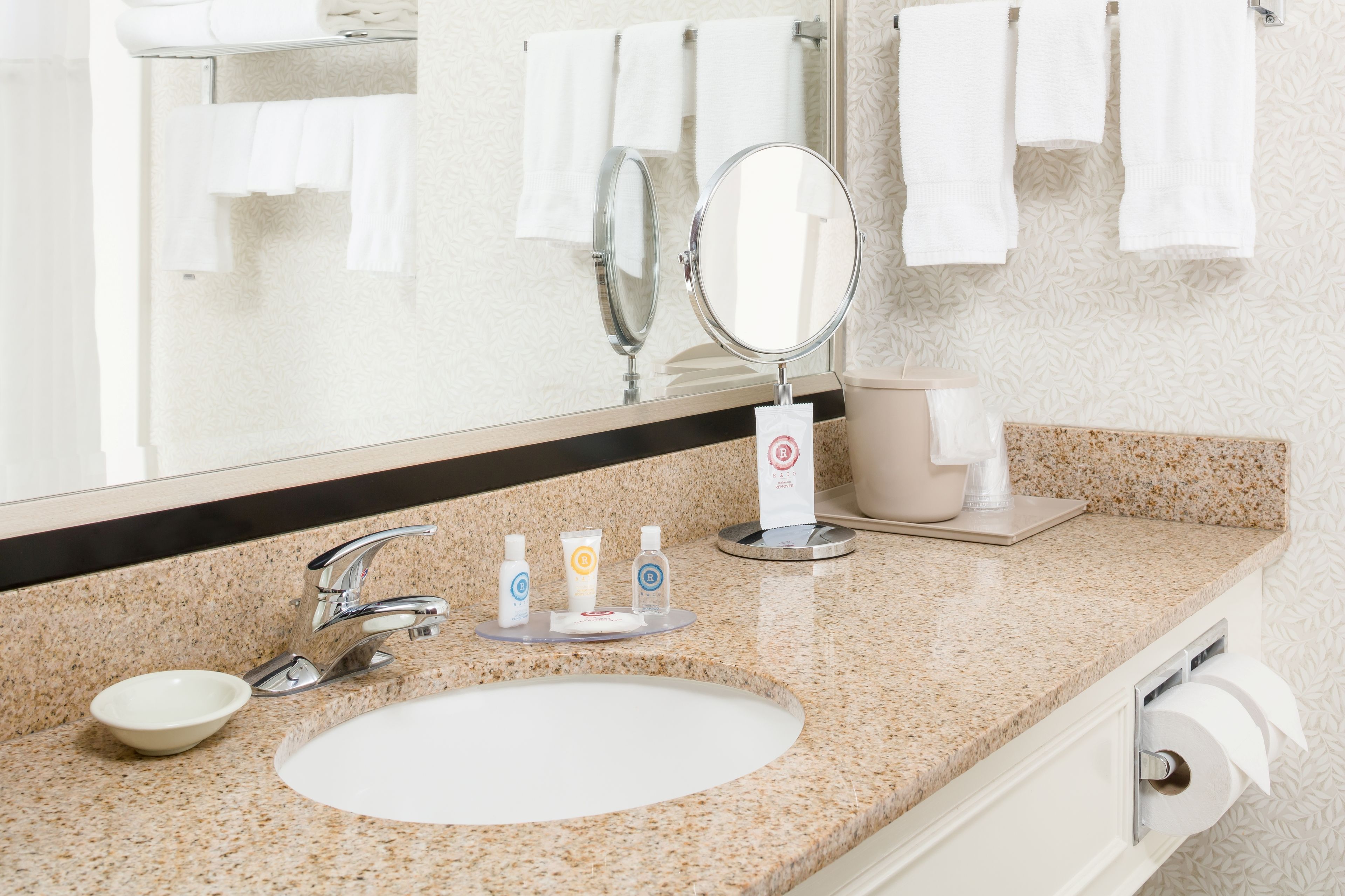 Un baño con encimera de granito, un lavabo blanco, un espejo y un perchero con varias toallas blancas.